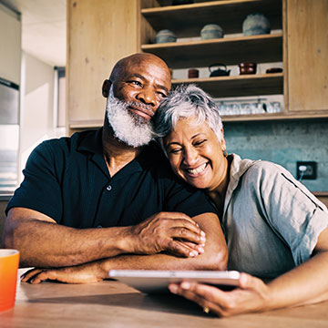 A married couple lean on each other as they laugh about what they saw on their tablet device. Links to Gifts of Real Estate